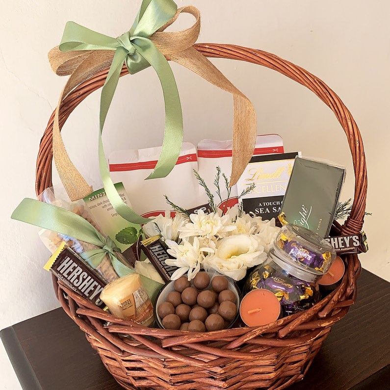 Wicker gift basket filled with chocolates, candles, coffee, flowers, and snacks